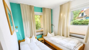 Une chambre avec deux lits simples, une table de chevet en bois, un miroir et une fenêtre avec des rideaux.