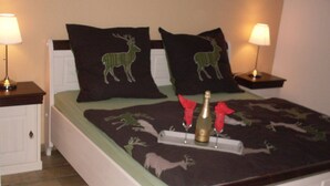 A bed with a green and black deer-themed bedspread, two matching pillows, a bottle of champagne, and two red napkins on a tray. - House Little Maddi Comfortable Vacation Home (Dornum)