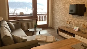 Ett vardagsrum med en soffa, ett soffbord, en tv som är monterad på en vägg av sten och en utsikt över ett snötäckt landskap genom stora fönster.