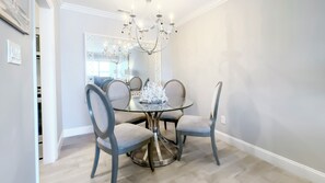 A dining area with a glass table, four grey upholstered chairs, a chandelier, and a decorative centerpiece. - 862 Ketch Court at The Sea Pines Resort (Hilton Head Island)