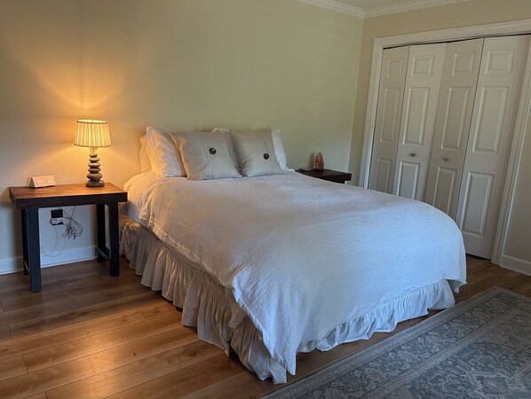 A neatly made bed with a white comforter and pillows, a bedside lamp, a small table with a clock, and a door in the background.