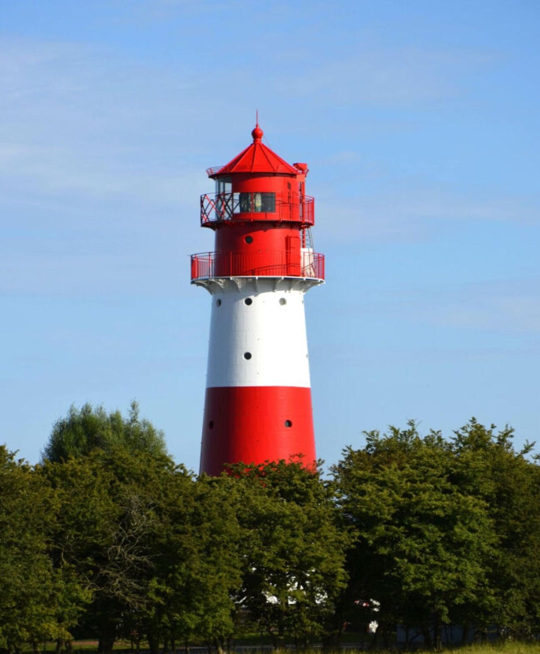 Un grand phare, avec une partie supérieure rouge et une partie inférieure blanche, entouré d’arbres.