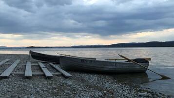 A rowboat with a paddle resting on a wooden dock, set on a pebbled shore with calm water and distant hills under a cloudy sky.