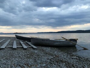 A rowboat with a paddle resting on a wooden dock, set on a pebbled shore with calm water and distant hills under a cloudy sky. - Holiday Apartment Seeloft (Konstanz)