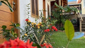 A building with a white facade, wooden shutters, and a small garden with red and yellow flowers. - Pousada Sempre Viva  (São João Batista do Glória)