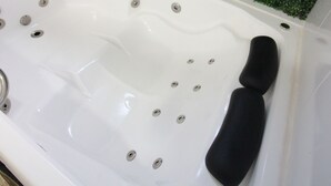 A white bathtub with multiple faucets, a black seat, and a green wall with a small shelf holding a potted plant and a picture frame. - Pousada Sempre Viva  (São João Batista do Glória)