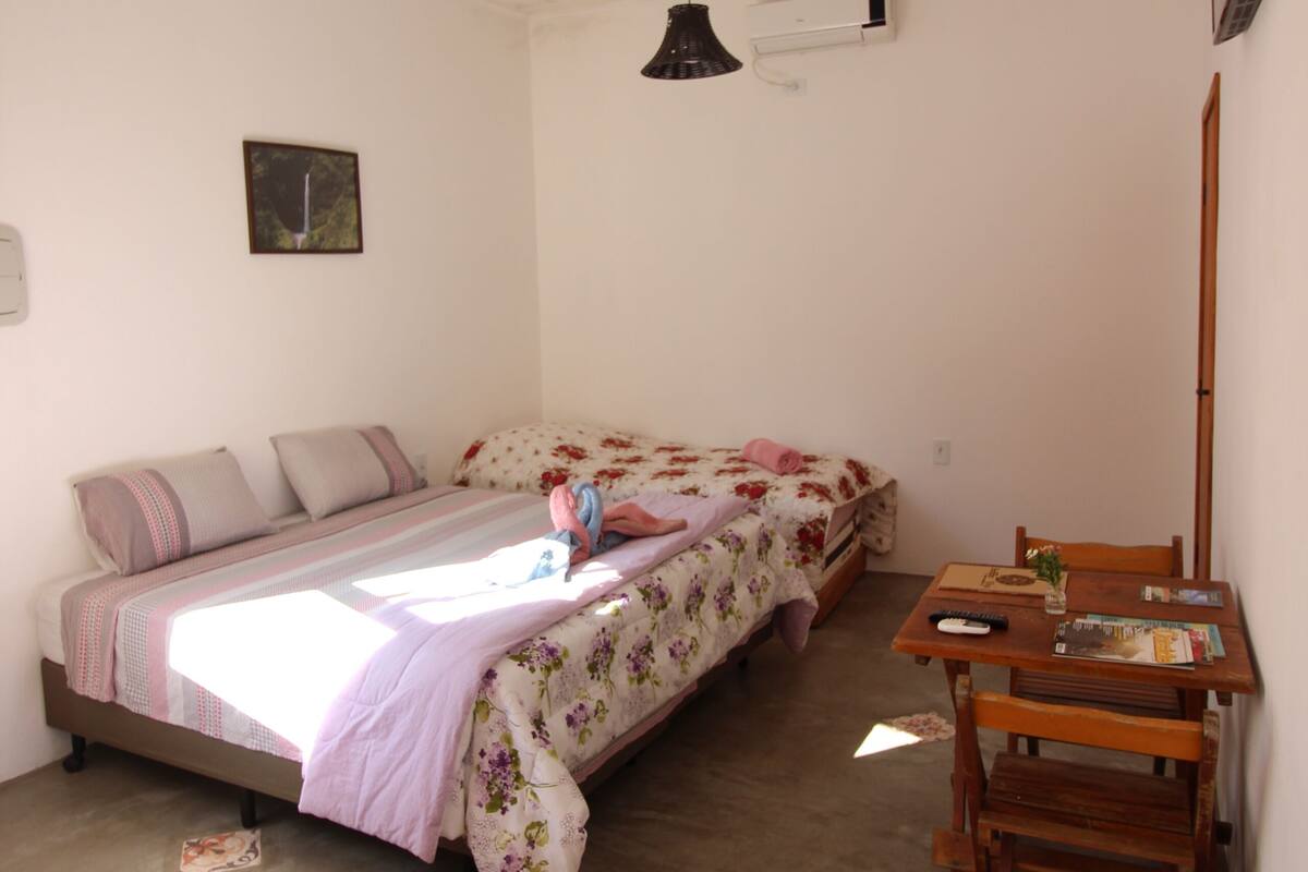 Una habitación pequeña con cama individual, una mesa de madera y sillas, una lámpara colgante y un televisor fijado a la pared.