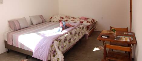 A small, single-bed room with a wooden table and chairs, a hanging light fixture, and a wall-mounted TV.