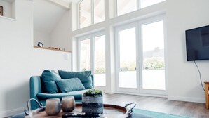 A modern living room with a teal sofa, a wooden coffee table with remote controls, and a large window offering a view of the outdoors. - Loheide Modern Retreat (Langenhorn)
