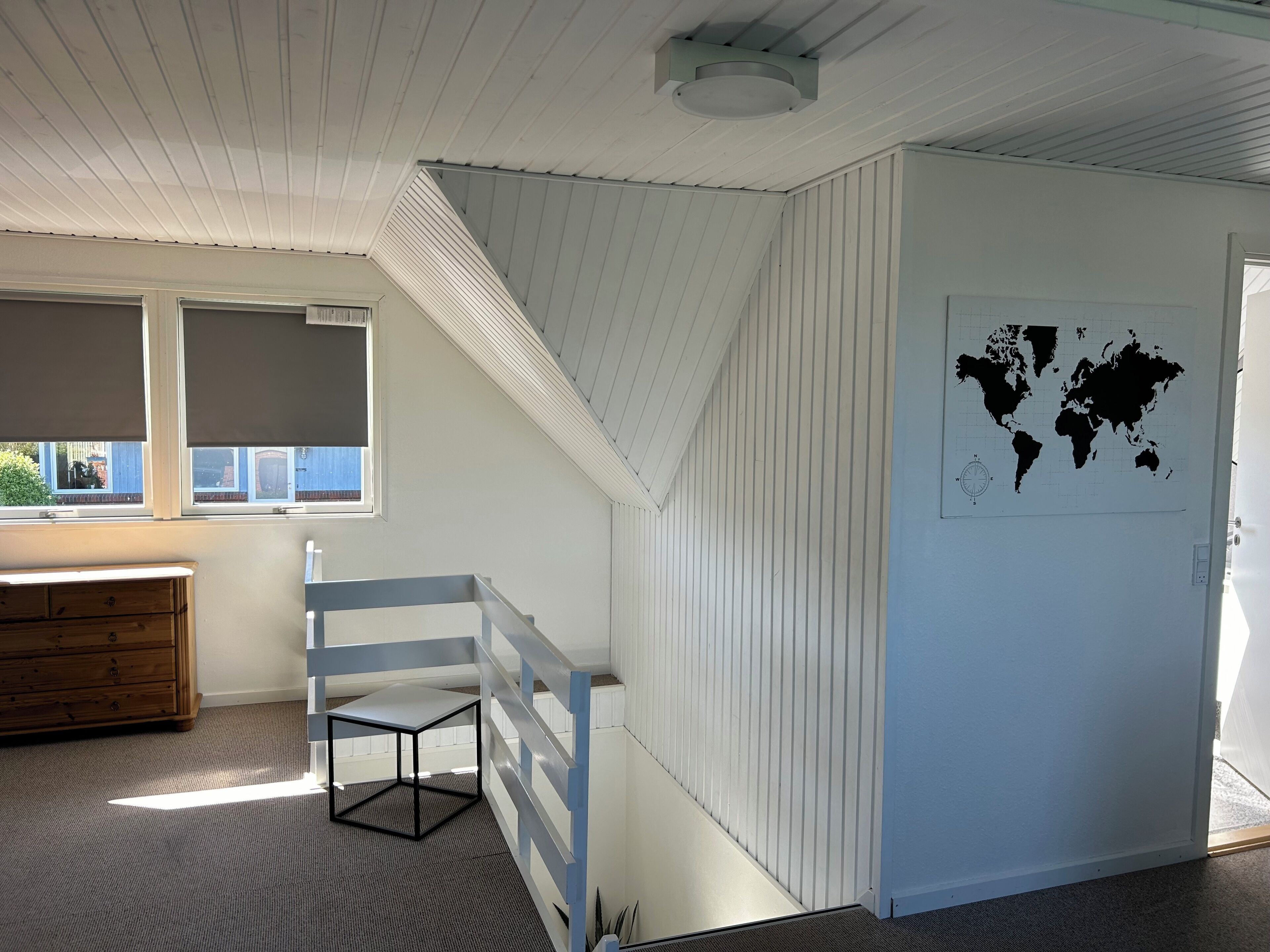 Une pièce avec un plafond en pente, une carte du monde accrochée au mur, une fenêtre avec des stores, une commode en bois et une petite table.