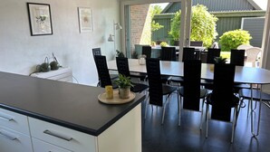 A modern kitchen with a dining area. There are two framed pictures on the wall, a small plant on the counter, and a window with a view of greenery outside.