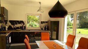 A modern kitchen with a large window, a dining area with orange chairs, and a pendant light.