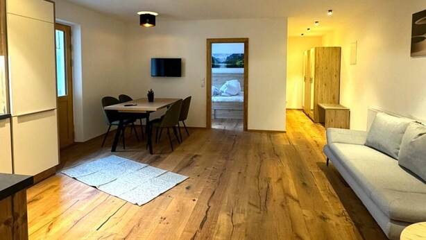 A modern living room with a wooden floor, a sofa, a dining table with chairs, a TV mounted on the wall, and a view of a lake through an open door.