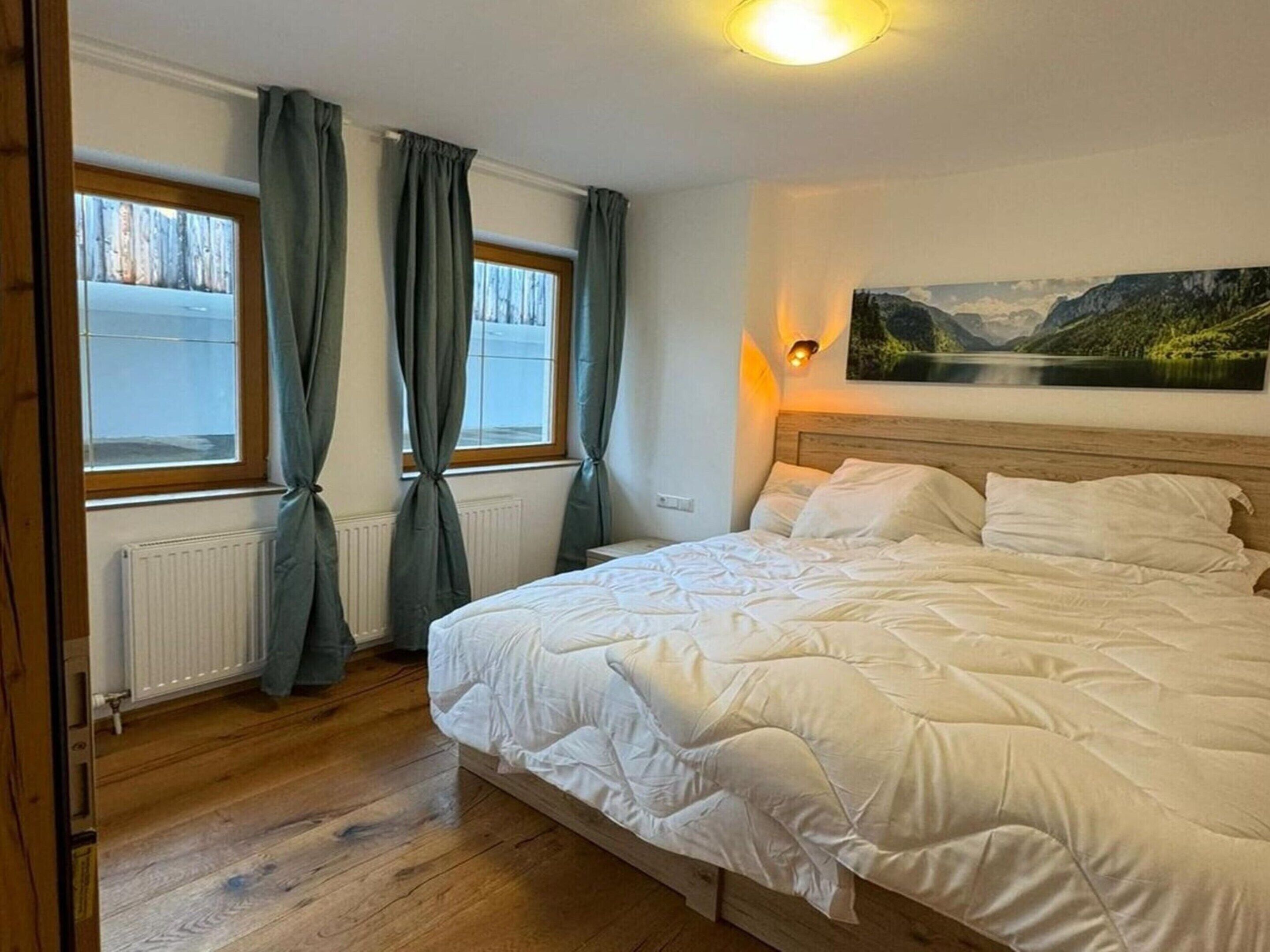 A bedroom with a large bed, two windows with curtains, a radiator, and a painting on the wall.