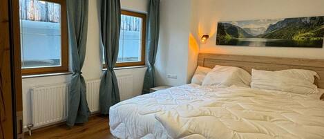 A bedroom with a large bed, two windows with curtains, a radiator, and a painting on the wall.