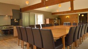 Un vaste espace dédié à la salle à manger, avec une longue table en bois entourée de chaises, un coin cuisine équipé d’appareils modernes et des poutres en bois au plafond.