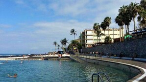 A swimming pool with a curved edge, surrounded by a stone wall and palm trees. - Belvilla by OYO Apartamento en Playa Las Carmenes (San Cristóbal de La Laguna)