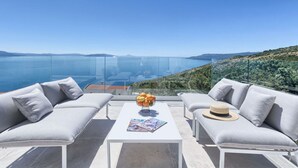 Una terraza con balaustrada de vidrio, asientos acolchados grises y una mesa con una revista y un frutero.