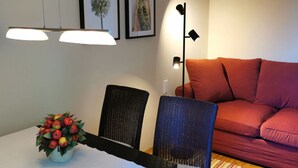 Ein Wohnzimmer mit einem roten Sofa, einem Esstisch mit Obst-Dekoration und zwei Wandleuchten.