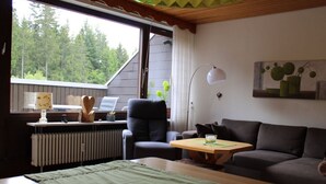 A cozy living room with a sofa, a wooden coffee table, a chair, and a desk with a lamp. A radiator is visible, and there is a window with a view of trees. - Holiday Apartment Biermann (Schluchsee)