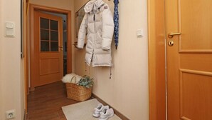 Um corredor com porta de madeira, um casaco branco pendurado em um gancho, um cesto com uma planta e um par de sapatos em um tapete.