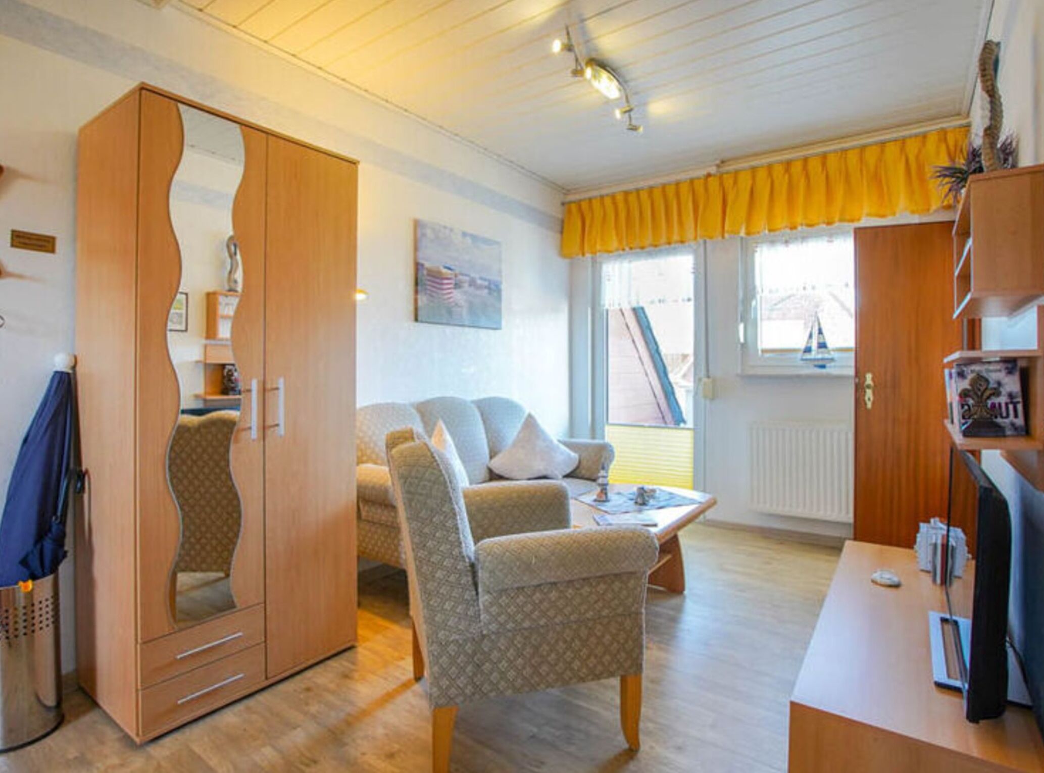 A cozy room with a wooden wardrobe, a patterned armchair, a small table, and a window with yellow curtains.
