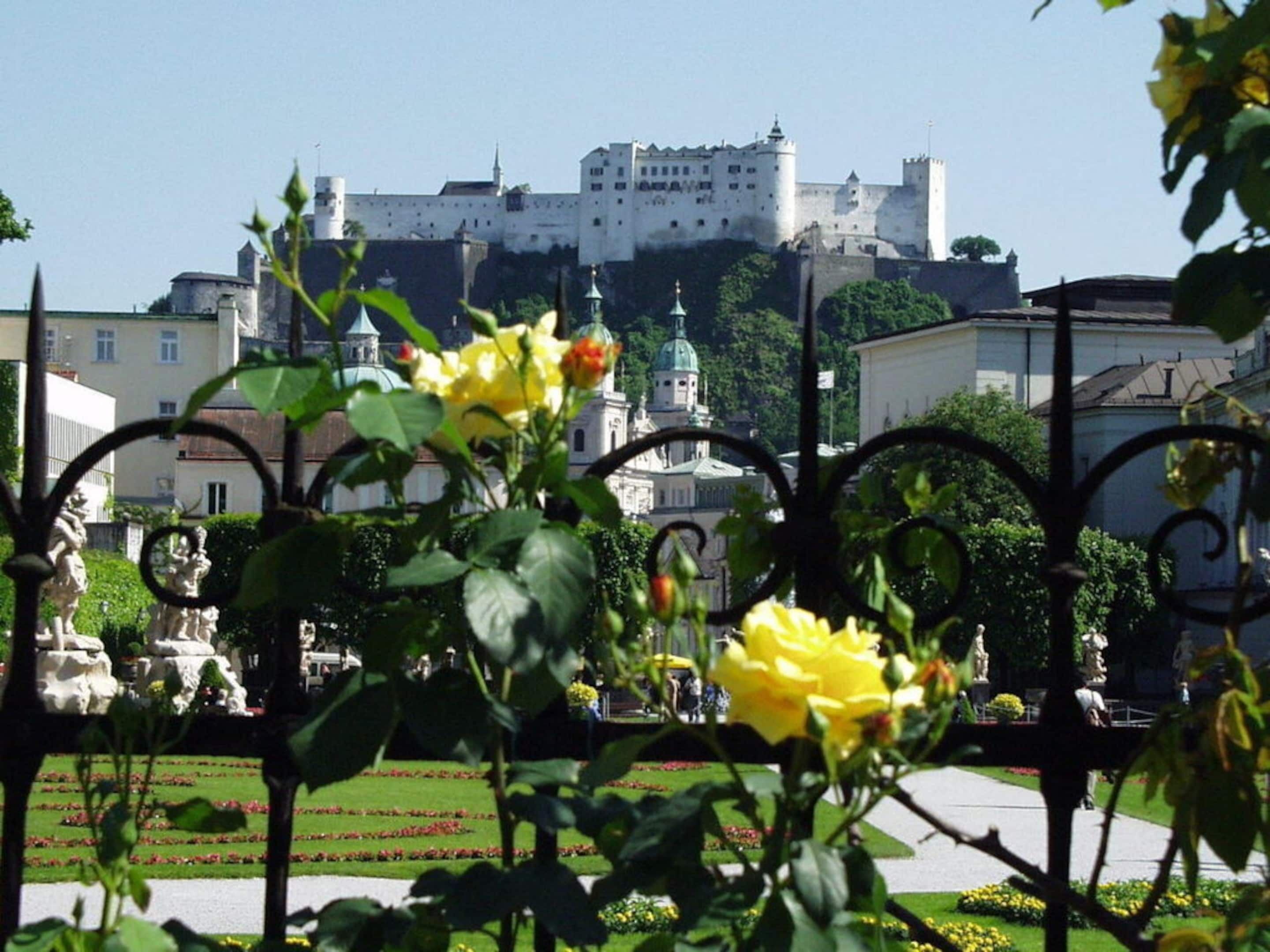 A castle with a fortified wall and a dome, surrounded by a well-maintained garden with yellow flowers and statues.
