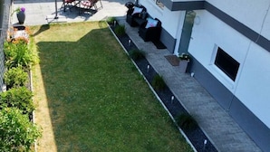 Un edificio moderno con un patio attrezzato con tavolo e sedie, un piccolo giardino con piante e un vialetto che conduce all'ingresso.