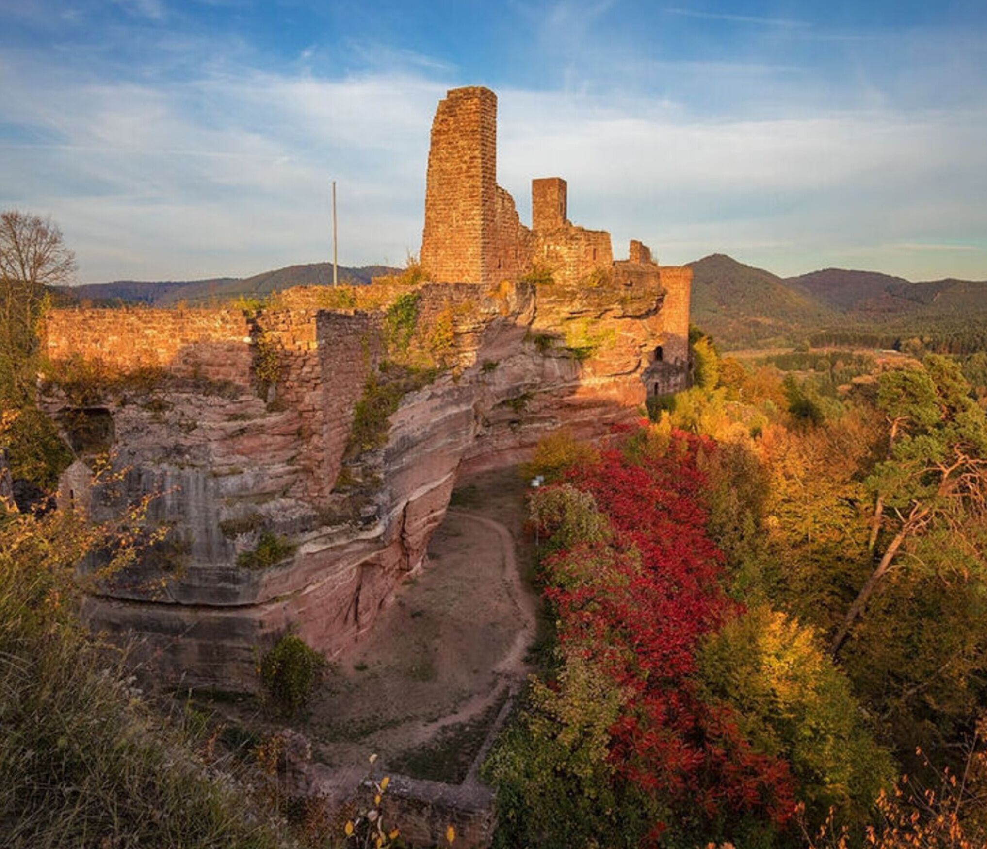 Un castillo de piedra histórico con ruinas, rodeado de follaje otoñal y colinas al fondo.