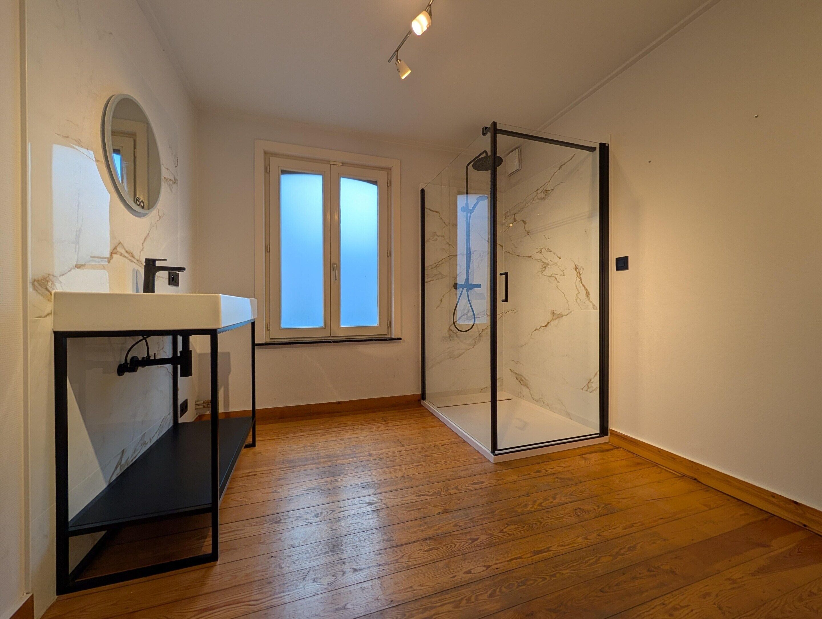 Ein modernes Badezimmer mit Duschkabine aus Glas, einem Waschbecken mit Marmorplatte und einem Fenster, das für natürliches Licht sorgt.