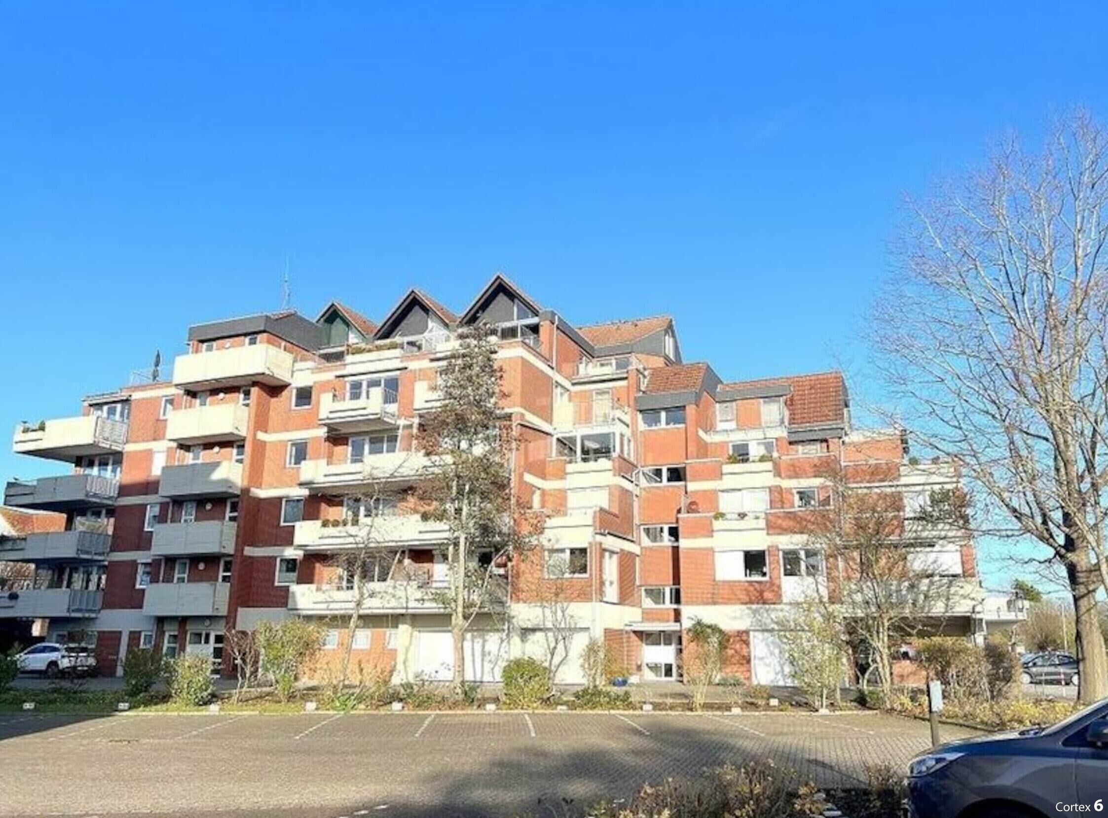 A multi-story residential building with a brick facade, balconies, and a parking area in front.