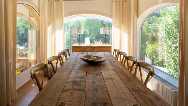 Une longue table à manger en bois, entourée de chaises, avec un grand saladier au centre, et une vue sur la verdure grâce à de grandes fenêtres.