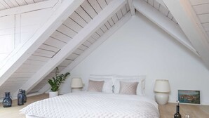 A bedroom with a sloped ceiling, a bed with white bedding, two bedside lamps, a small table with a bottle and glasses, and a potted plant. - Secret Apartments - One Bedroom Apartment (Dubrovnik)