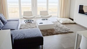 Uma sala de estar moderna com um sofá cinzento, duas poltronas brancas, uma mesa de centro e uma TV na parede. Grandes janelas proporcionam vista para a praia.