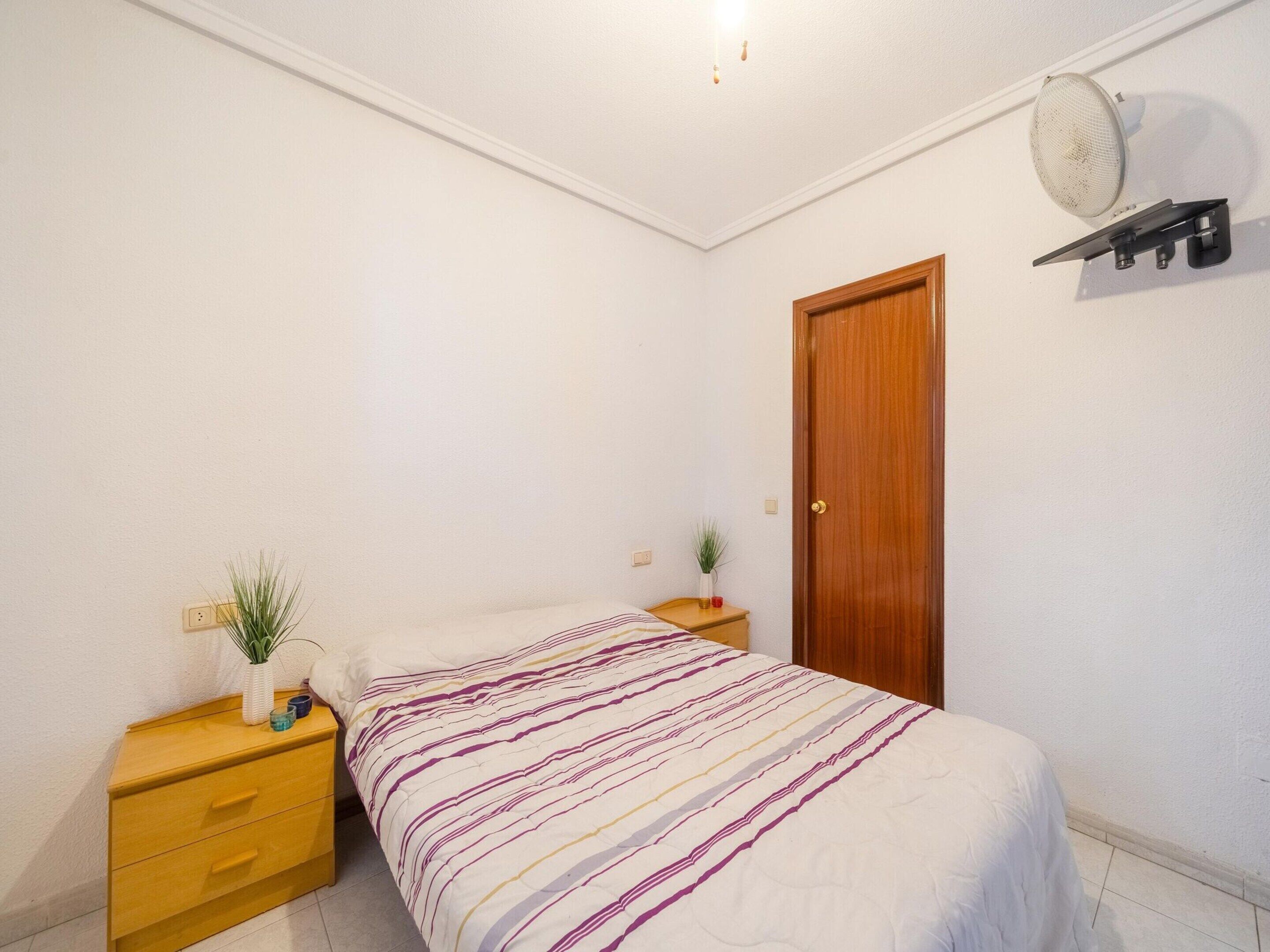 Una camera da letto con un letto, un comodino in legno, un tavolino con una pianta, una ventola a muro e una porta in legno.