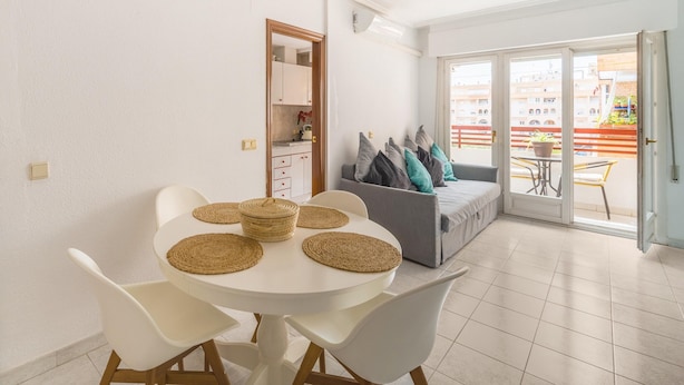 A dining area with a white table and chairs, a grey sofa with cushions, and a balcony with a view of buildings.