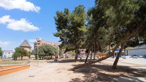 A sandy plaza with trees, a stone structure, and buildings in the background.