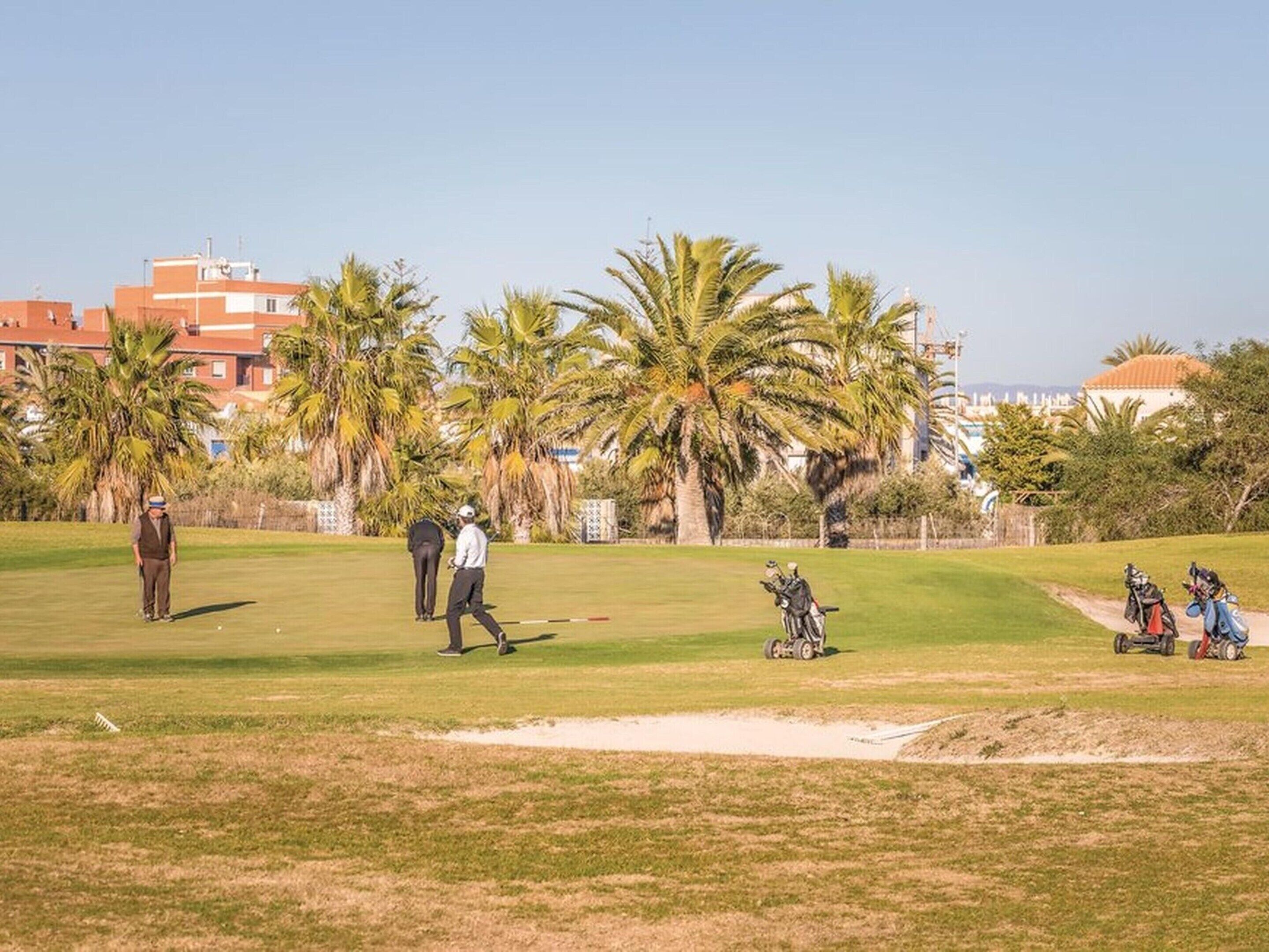 A golf course with a sand bunker, palm trees, and buildings in the background.