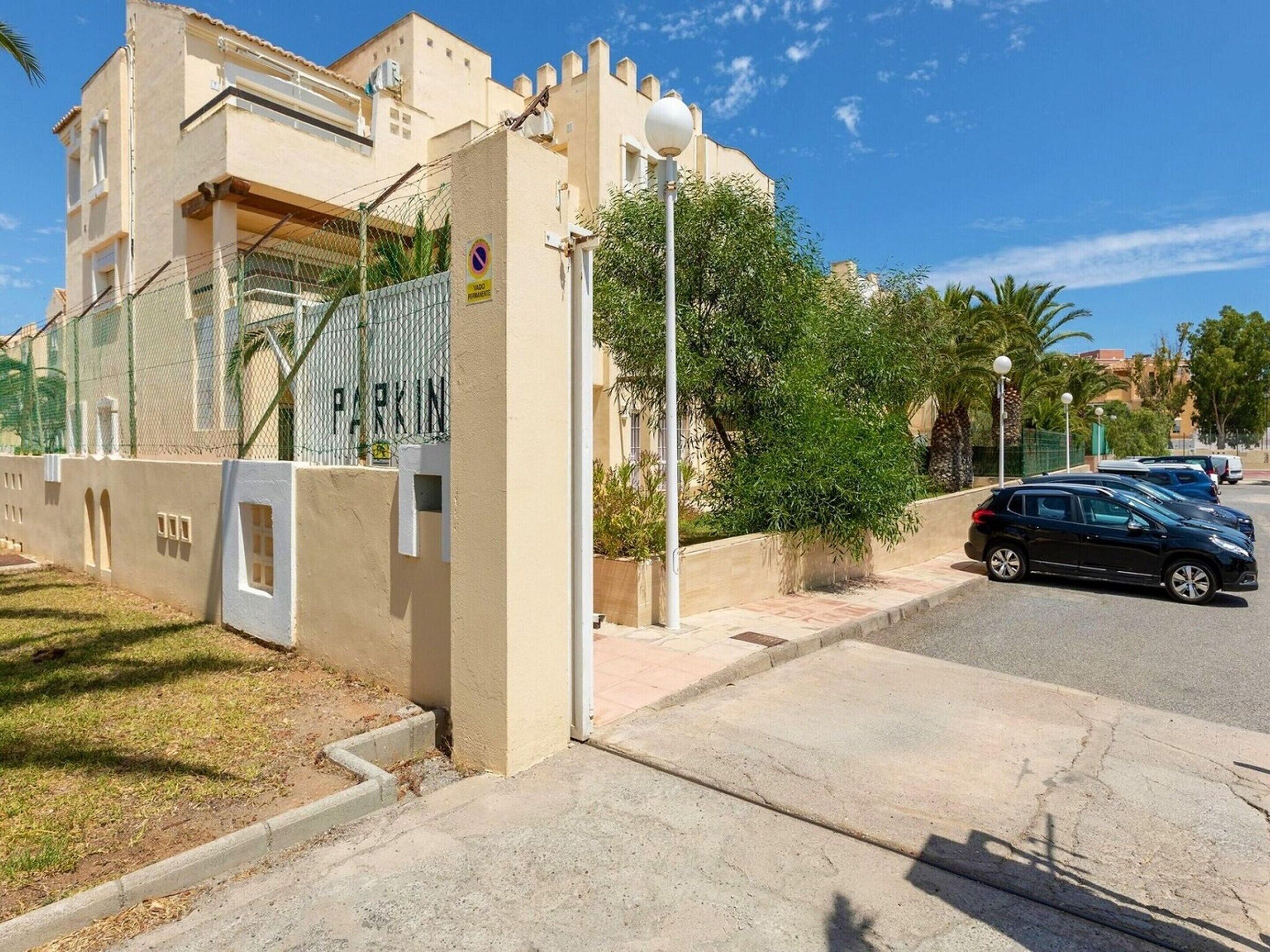 A residential building with a gated entrance, a parking area, and a street with parked cars.