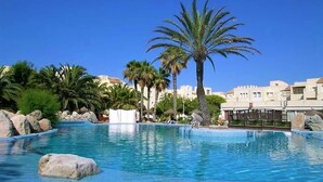 A swimming pool with clear blue water, surrounded by rocks and palm trees, with buildings in the background. - Belvilla by OYO Macael (Roquetas de Mar)