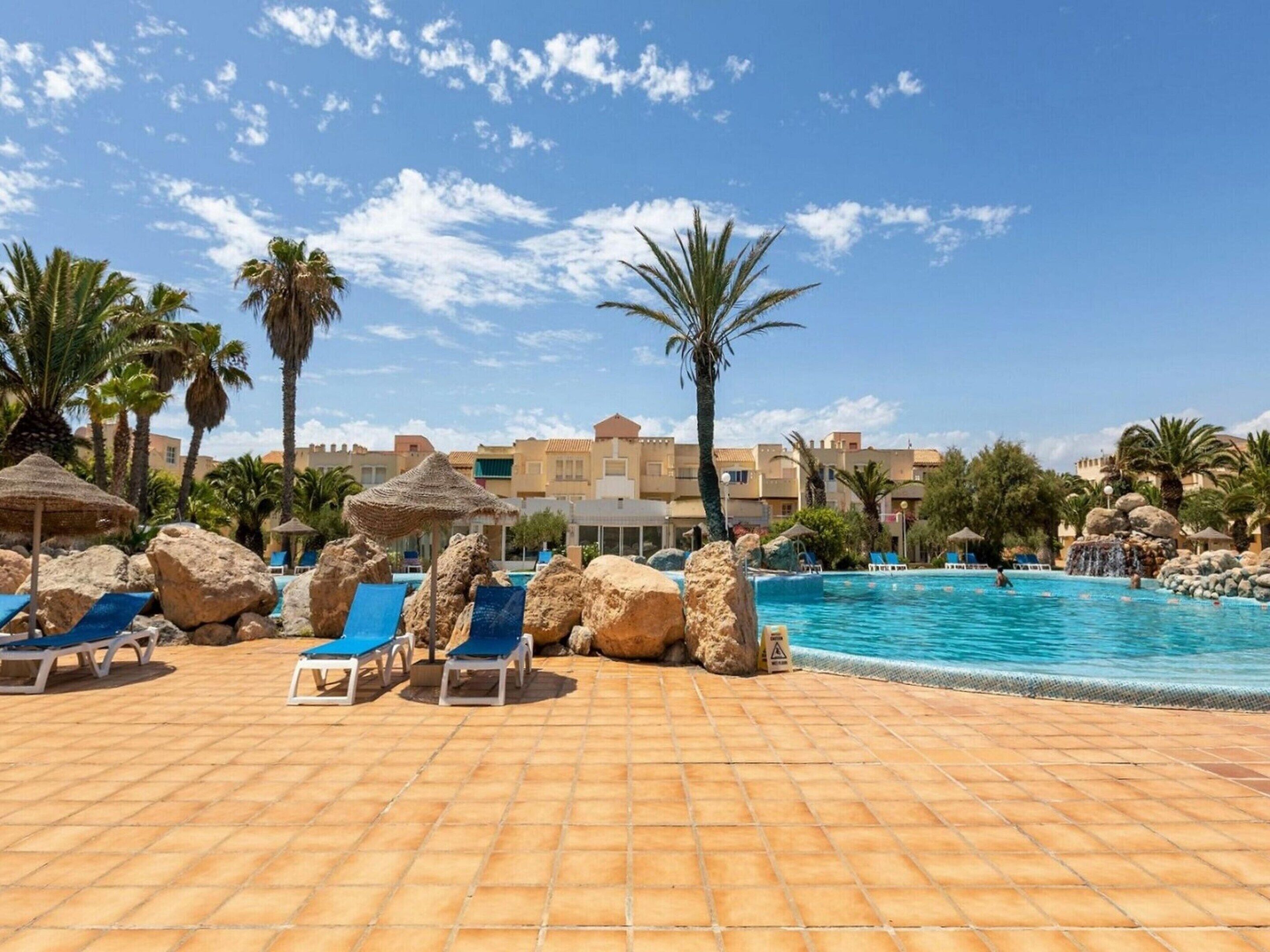 A pool area with lounge chairs and rocks, palm trees, and buildings in the background.