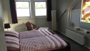 A bedroom with a bed, two windows, a chair, a radiator, and a view of a wooden structure outside. - Seagull Modern Retreat (Wangerland)