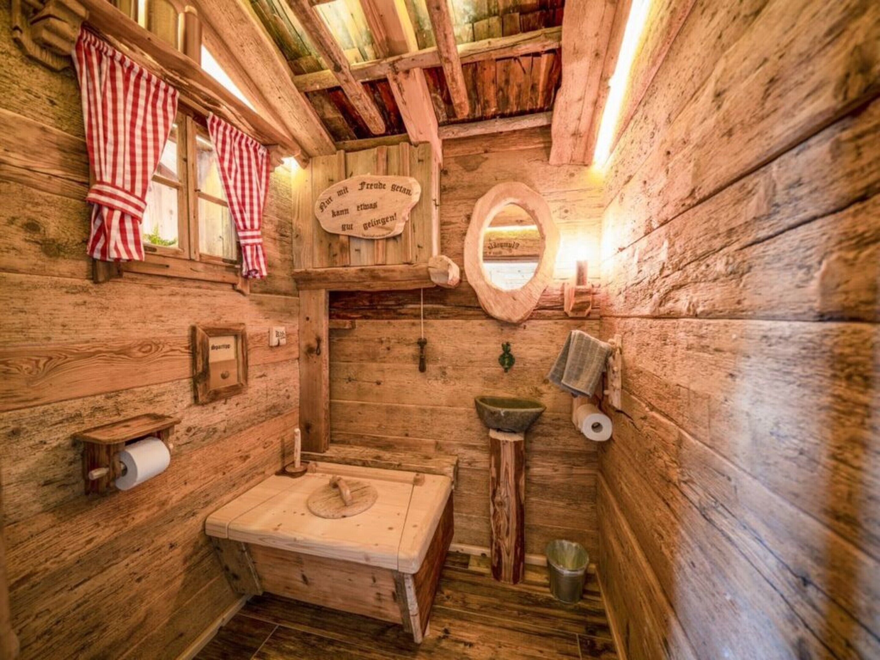 Ein rustikales Badezimmer einer Holzhütte mit einer hölzernen Bank, einer Toilette aus Holz, einem Waschbecken und einem Spiegel.