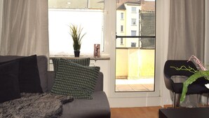 A living room with a sofa, a chair, a potted plant, and a view of buildings through a window. - Cosy Apartment in Oberhausen With Balcony (Oberhausen)