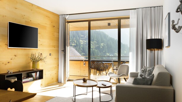 Un soggiorno moderno con arredamento in legno, una televisione a schermo piatto, un divano, due tavolini e un balcone con vista sulle montagne.