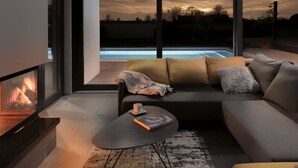 A modern living room with a sofa, coffee table, and fireplace. A large window offers a view of a swimming pool and sunset.