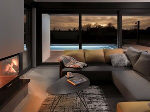A modern living room with a sofa, coffee table, and fireplace. A large window offers a view of a swimming pool and sunset. - Villa Linea Comfortable Holiday Residence (Fazana)