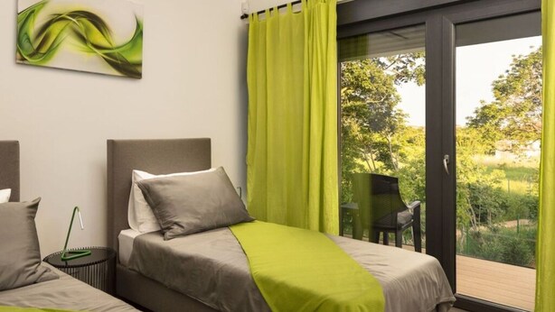 A modern bedroom with two beds, a green curtain, and a view of greenery outside.