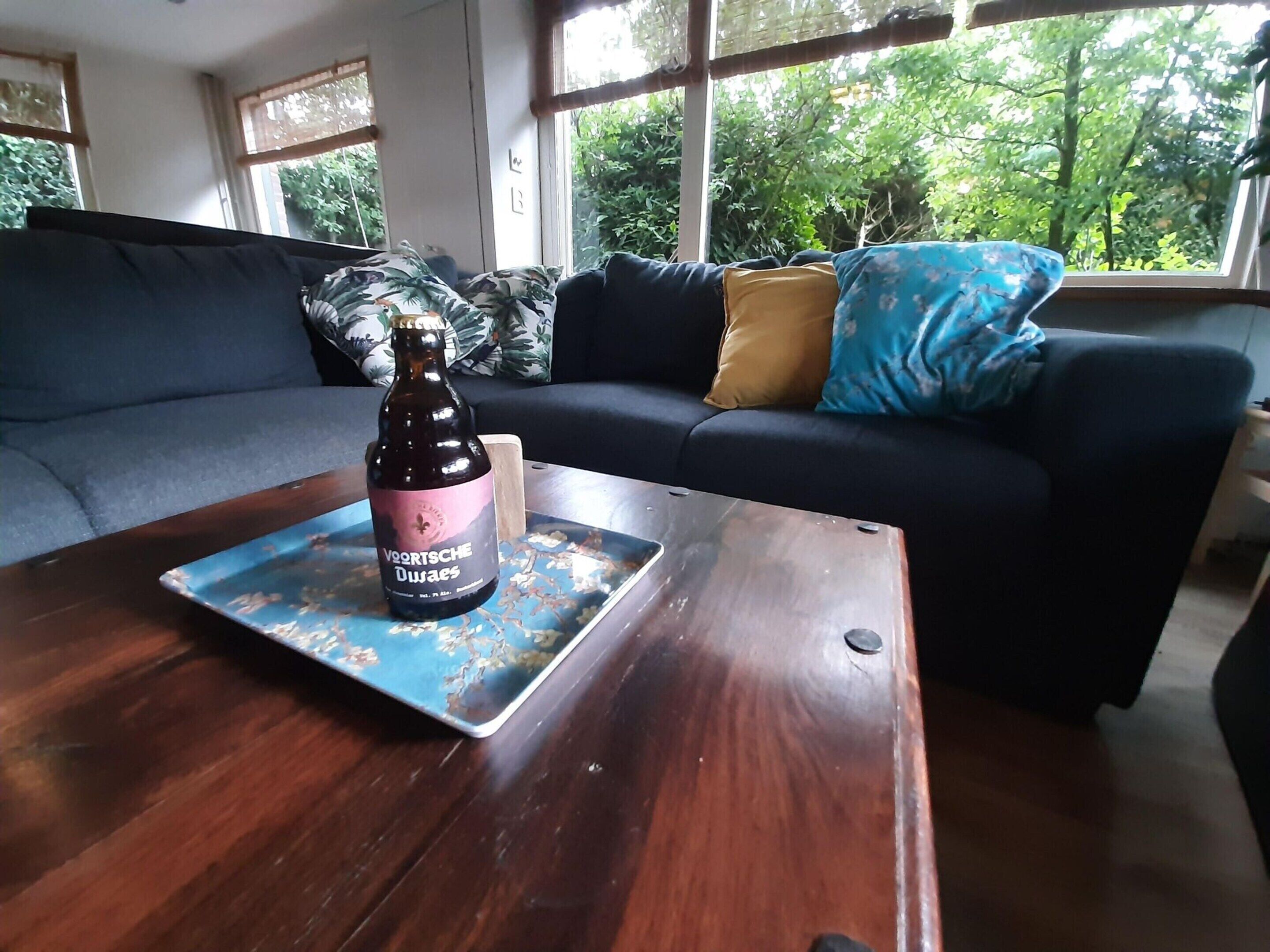 A room with a dark blue sofa, a wooden coffee table with a bottle of beer, and a view of greenery through large windows.