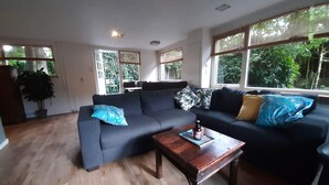 A modern living room with a dark blue sectional sofa, a wooden coffee table, and large windows offering a view of greenery outside. - Holiday Home in Voorthuizen With Hot Tub (Voorthuizen)